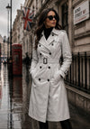 Woman in a white trench coat standing on a rain-soaked street with a British flag and sign in the background.